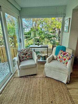 Ein Zimmer mit zwei Sesseln, einem kleinen Tisch und Blick auf einen Garten durch große Fenster.