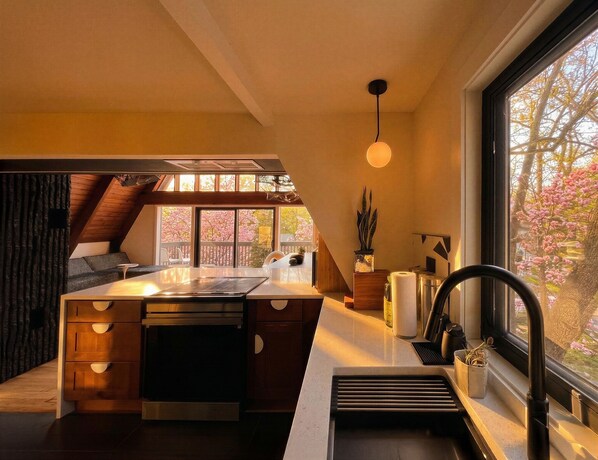 A modern kitchen with a central island, a built-in oven, and a window with a view of cherry blossoms. - Custom A-Frame | Lake Access • Fire Pit (Highland Lakes)