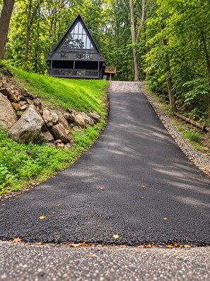 A cabin with a triangular roof, surrounded by a forested area and a paved road leading to it.