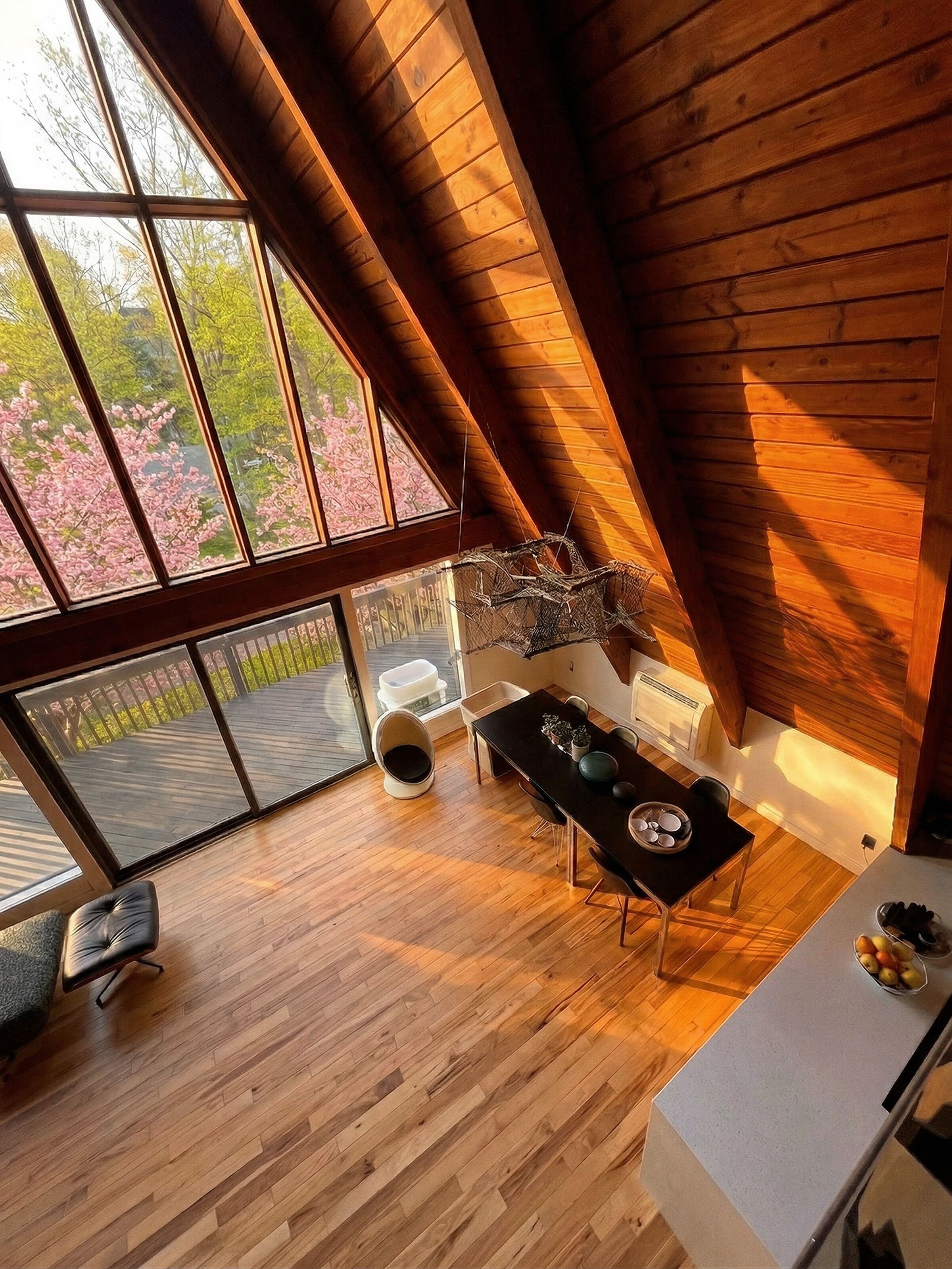 A modern loft with a high ceiling, wooden beams, a dining area with a table and chairs, and large windows offering a view of cherry blossoms.