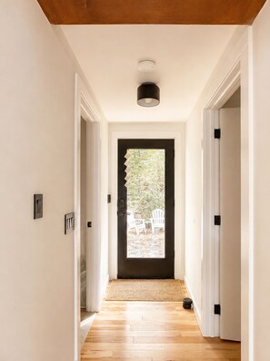 A hallway with a wooden floor leading to a door with a view of outdoor chairs.