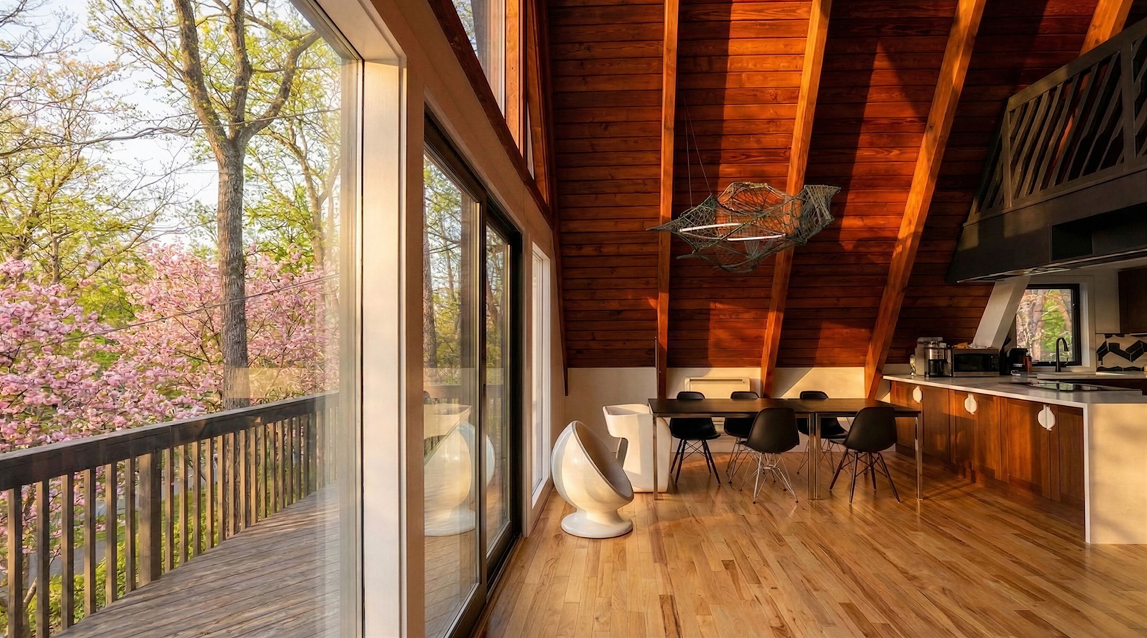 A modern kitchen with wooden beams, a dining area with chairs, and large windows overlooking a garden with pink flowers.