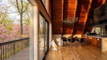 A modern kitchen with wooden beams, a dining area with chairs, and large windows overlooking a garden with pink flowers.