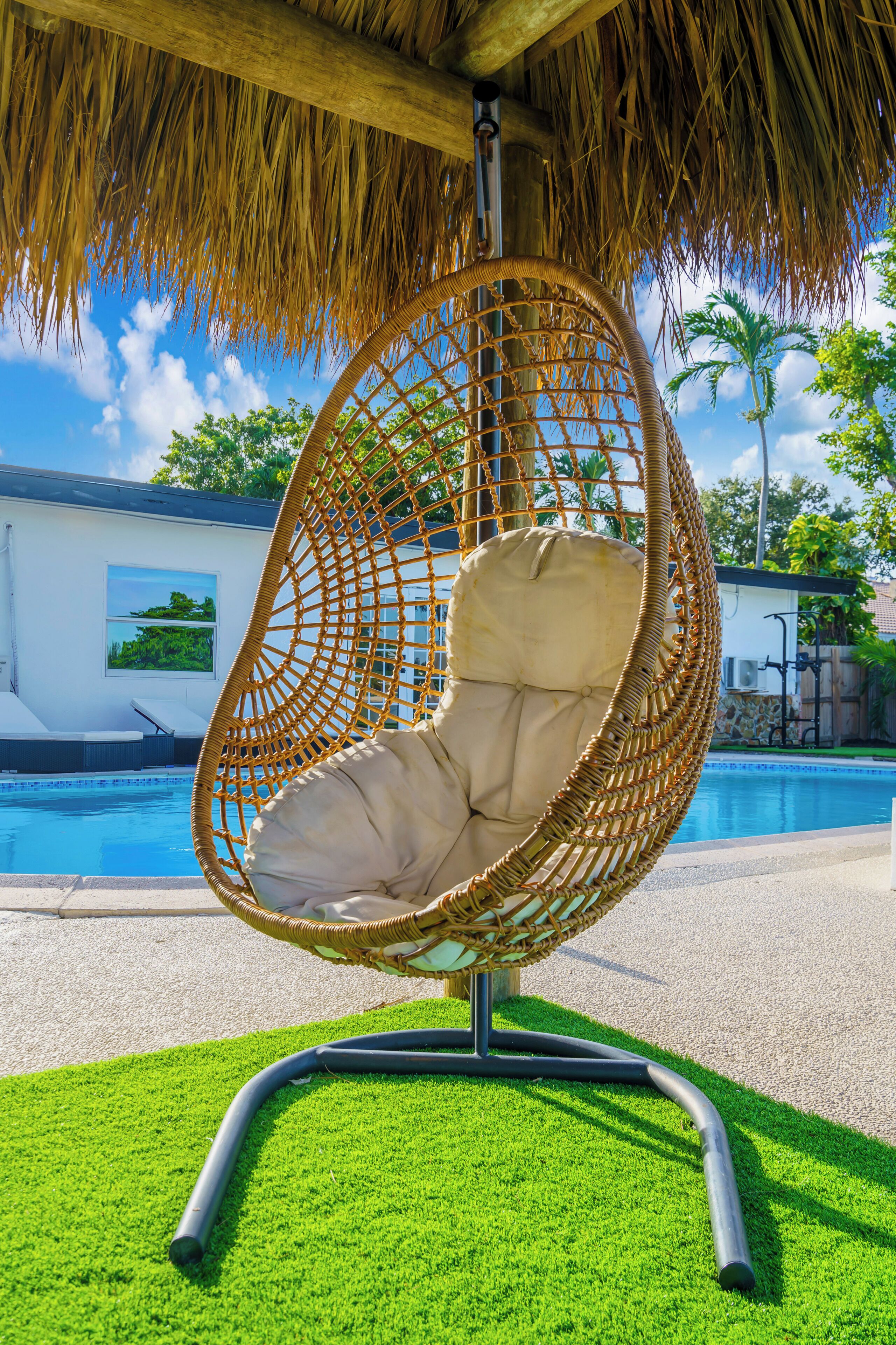 A hanging chair with a cushion by a poolside, a thatched roof overhead, and a clear view of the pool and surrounding greenery.