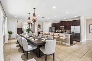 A spacious dining area with a long table and multiple chairs, a kitchen with dark cabinets and stainless steel appliances, and a chandelier overhead. - PacMan, Foosball, Shuffleboard, Large Backyard, Chef Kitchen: Metropolis Estate (Kissimmee)