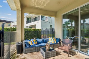 A covered outdoor seating area with a sofa and chairs, a trash bin, and a glass door leading to a balcony. - PacMan, Foosball, Shuffleboard, Large Backyard, Chef Kitchen: Metropolis Estate (Kissimmee)