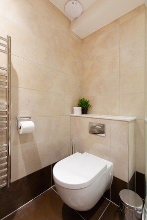 A modern bathroom with a toilet, a towel rack, a small shelf with a plant, and a trash bin. - Lovely 1BR Fitzrovia - Soho (London)