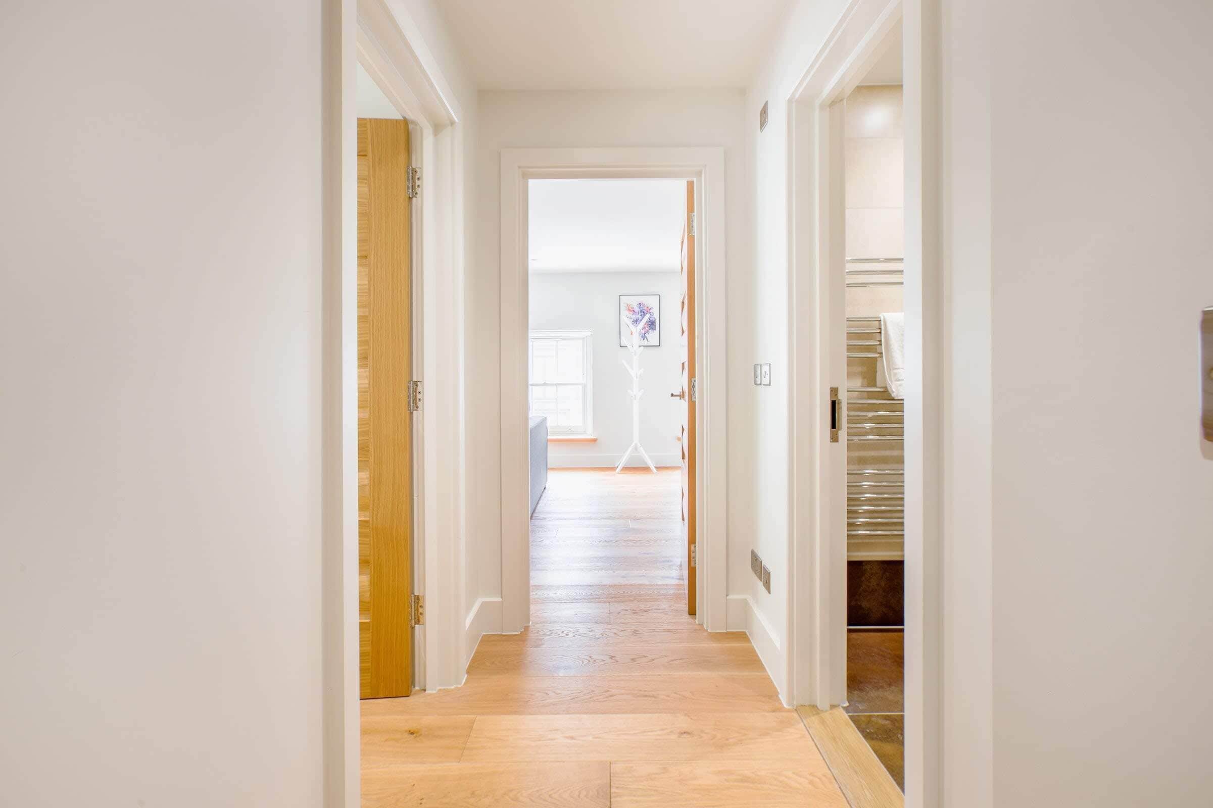 A hallway with wooden flooring, white walls, and a door leading to a room with a window.