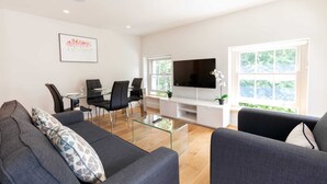 Ein modernes Wohnzimmer mit einem Glastisch, einem wandmontierten Flachbildfernseher und einem Essbereich mit Glastisch und schwarzen Stühlen.