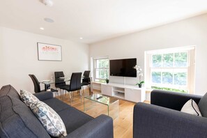 A modern living room with a glass coffee table, a flat-screen TV mounted on the wall, and a dining area with a glass table and black chairs. - Lovely 1BR Fitzrovia - Soho (London)