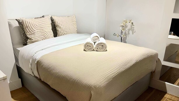 Ein ordentlich bezogenes Bett mit einer beigen Tagesdecke und Kissen, eine kleine Vase mit weißen Blumen und ein Holzboden.