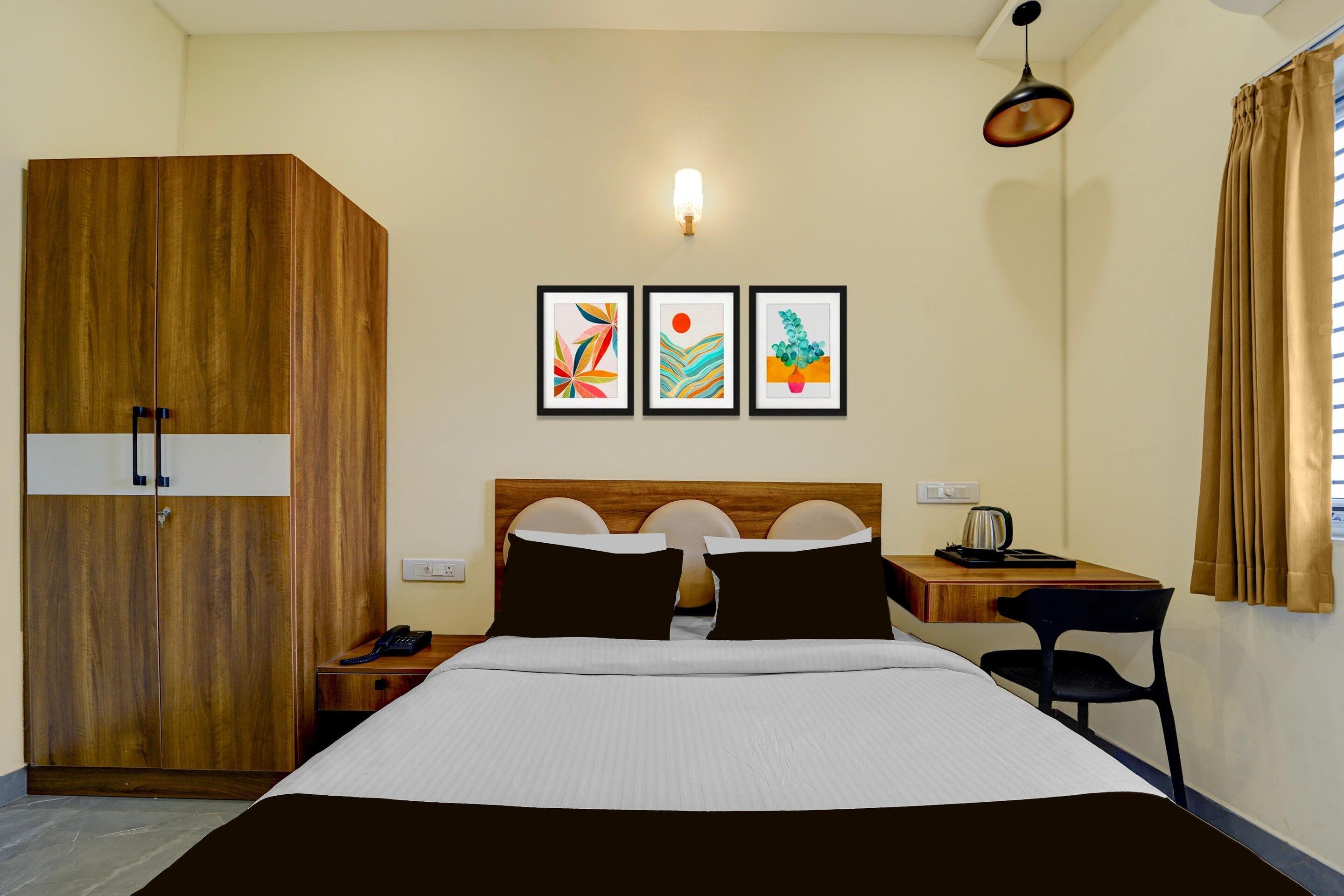 A hotel room with a bed, a desk, a wardrobe, and three framed artworks on the wall.