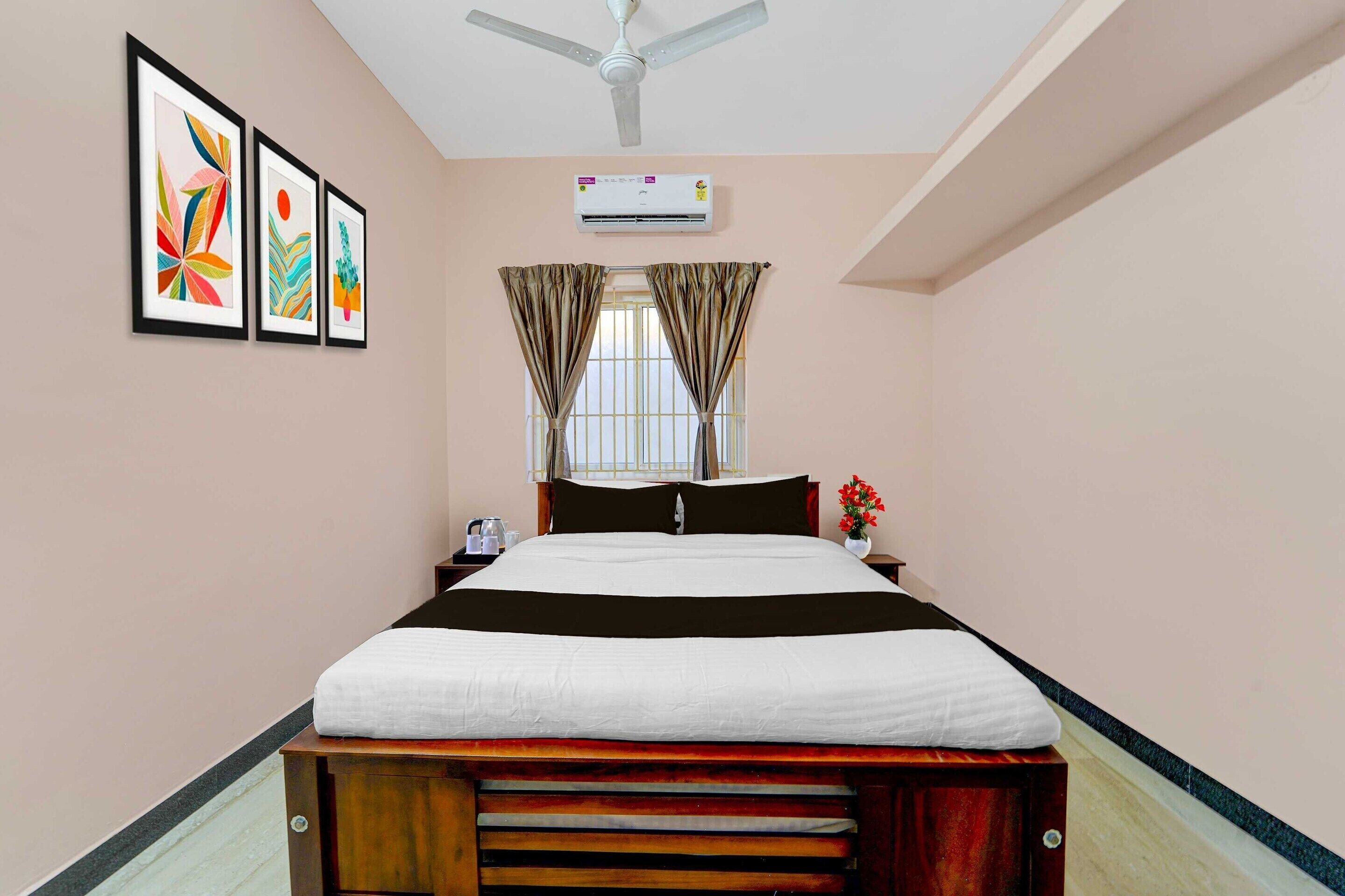 A hotel room with a bed, a ceiling fan, an air conditioning unit, a window with curtains, and framed artwork on the wall.