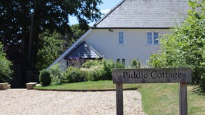 Un panneau indiquant « Piddle Cottage », avec un chemin de gravier menant à une maison dotée d’un toit en pente et d’un jardin en premier plan.