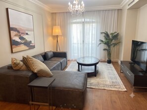 A modern living room with a sofa, a coffee table, a TV, a chandelier, and a large window with curtains. - 2-bedroom apartment Cape Royale Luxury Suite (Cape Town)