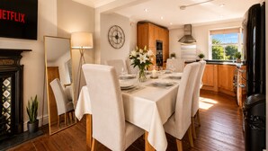 Uma sala de jantar com uma mesa posta para quatro pessoas, armários de madeira, um relógio na parede e uma cozinha ao fundo.