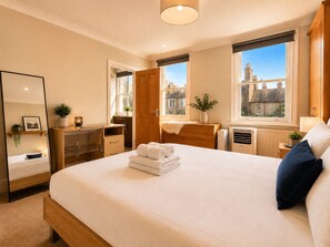 A hotel room with a bed, a desk, a dresser, a mirror, and a window with a city view. - Fitzrovia 2-Bed, 2-Bath Apartment | Near Tottenham Court Road (London)