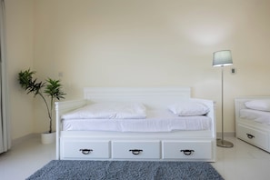 A white bed with a headboard, two bedside tables with drawers, a floor lamp, and two potted plants.