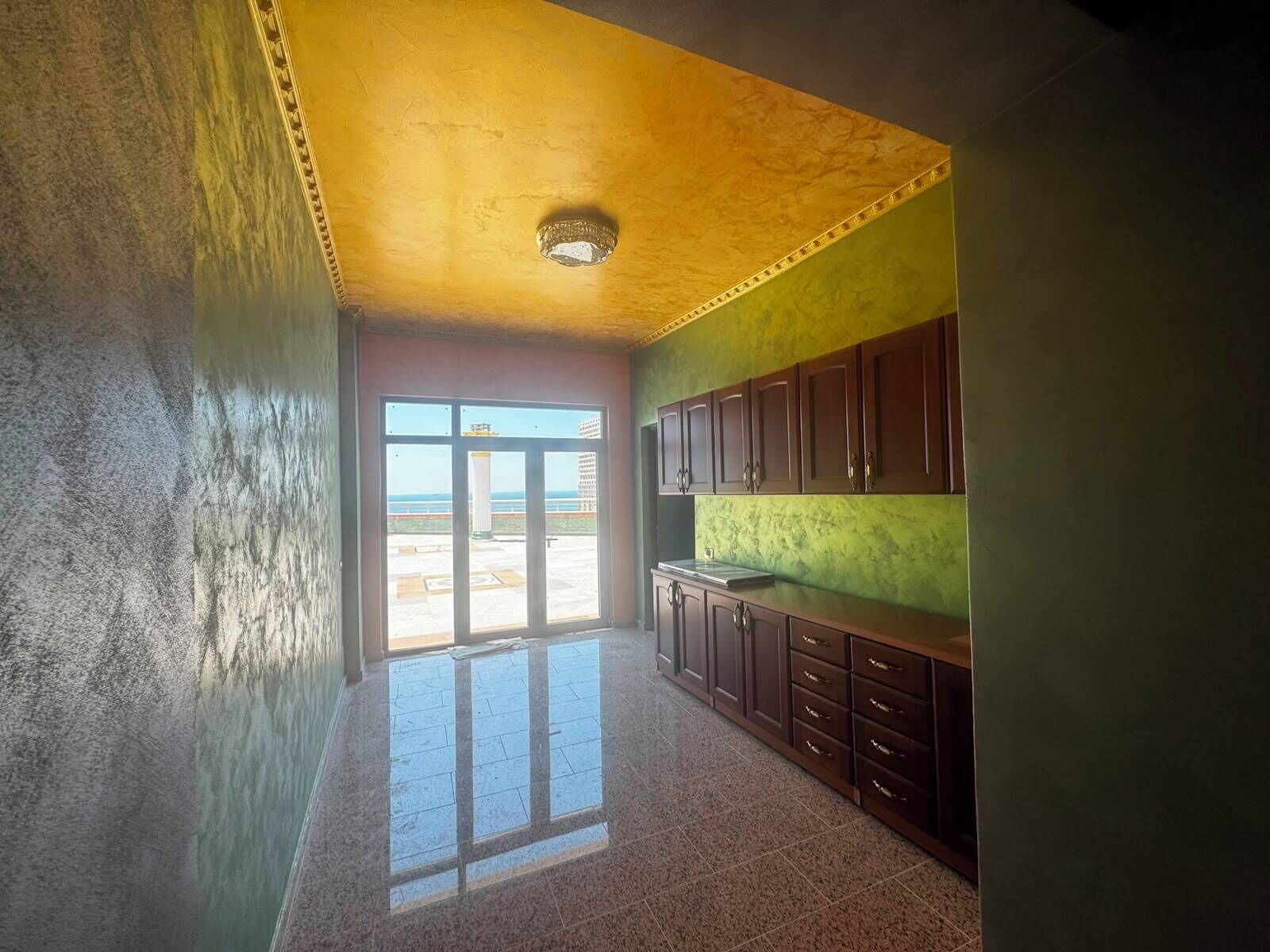 A room with a large sliding glass door leading to an outdoor area, a kitchen with dark wooden cabinets, and a textured yellow ceiling.