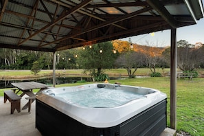 Un jacuzzi bajo una zona techada, con una mesa de picnic cerca, rodeado de un paisaje natural.