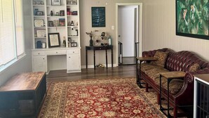 Ein Wohnzimmer mit einem großen roten und beigefarbenen Teppich, einem Ledersofa, einem Holztisch, einer weißen Regaleinheit mit diversen Gegenständen und einem Deckenventilator.
