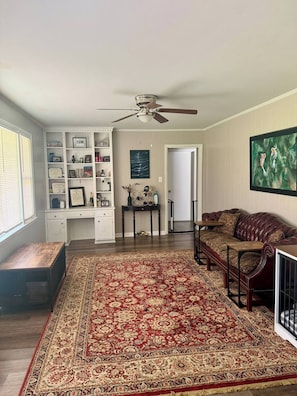 A living room with a large red and beige patterned rug, a leather sofa, a wooden coffee table, a white shelving unit with various items, and a ceiling fan. - Hillwood Circle home with fast EV charger (Augusta)