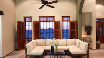 Una habitación con un sofá seccional color beige, una mesita y un ventilador de techo. Hay tres ventanas con contraventanas de madera, cada una con vistas al océano.