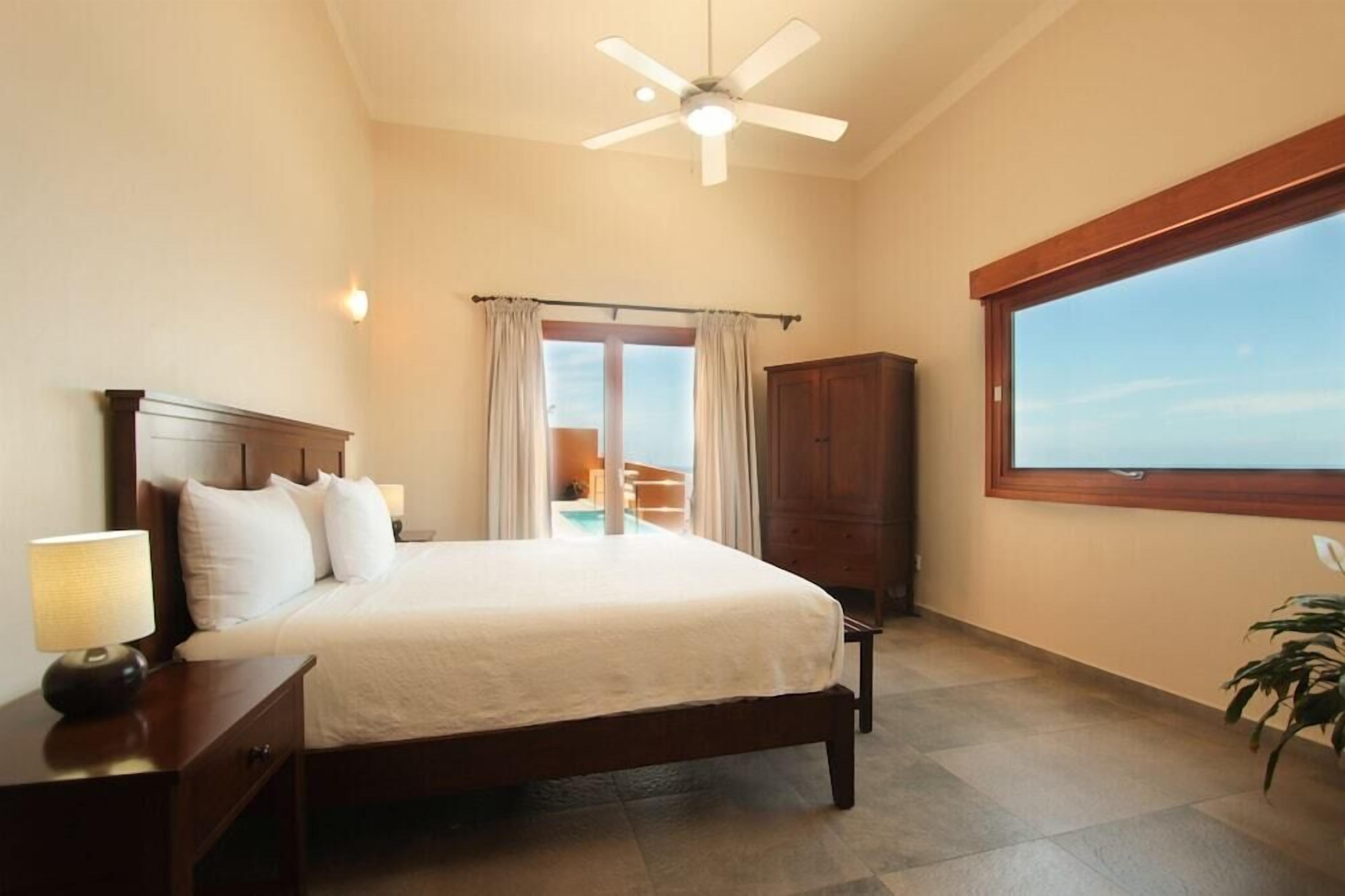 A bedroom with a bed, nightstand, lamp, ceiling fan, window, and a view of a balcony.
