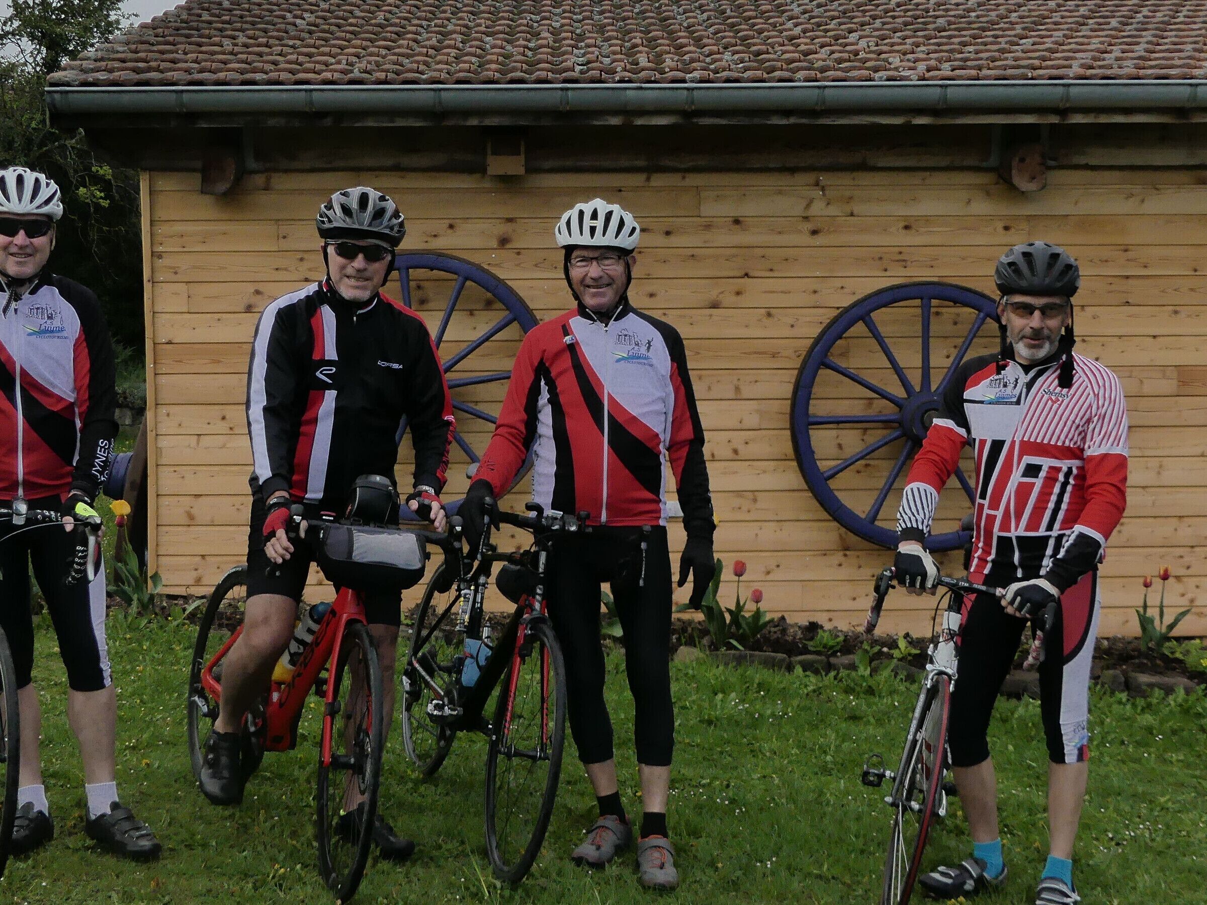 Four cyclists standing in front of a wooden building with a tiled roof.