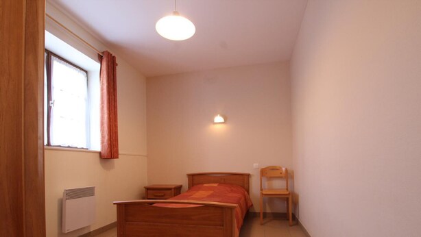 A bedroom with a wooden bed, a nightstand, a chair, a radiator, and a window with curtains. - Cozy house, pets allowed - 5 guests (Saint-Georges-de-Rouelley)