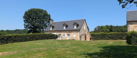 A stone house with a sloped roof, surrounded by a well-maintained lawn and a hedge.