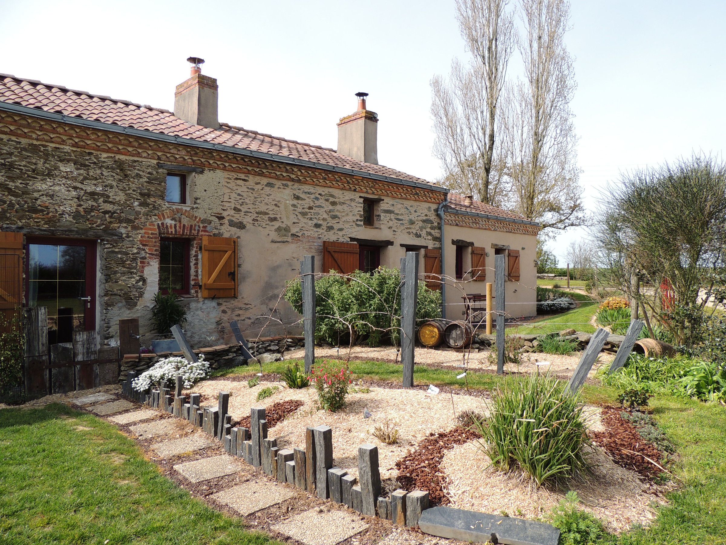 Una casa de piedra con techo de tejas, contraventanas de madera y un jardín con cerca de madera y plantas.