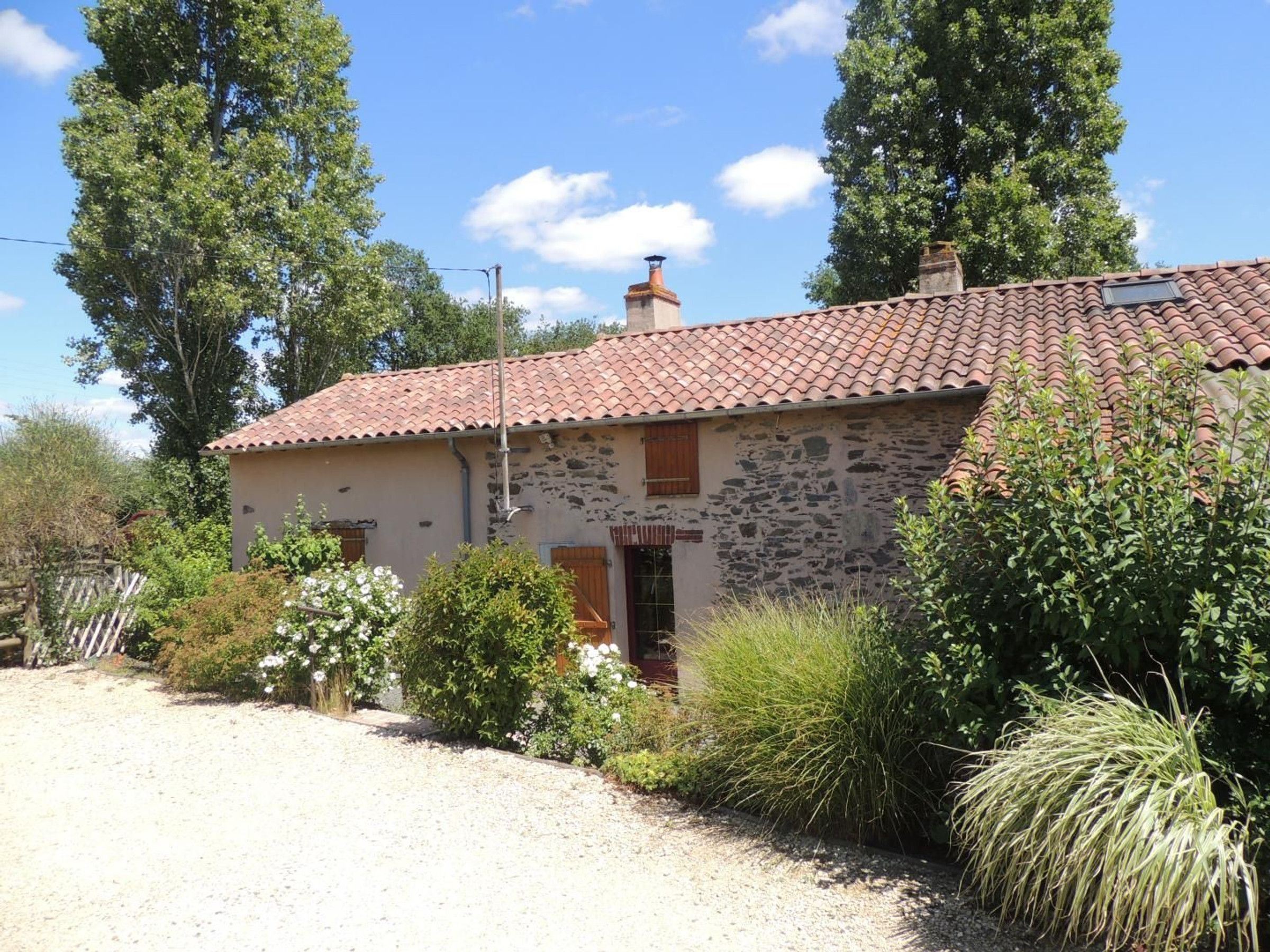 A rustic stone building with a tiled roof, surrounded by greenery and a gravel driveway.