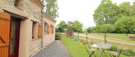 A rustic building with a gravel path leading to a table and chairs, surrounded by greenery and a fence.