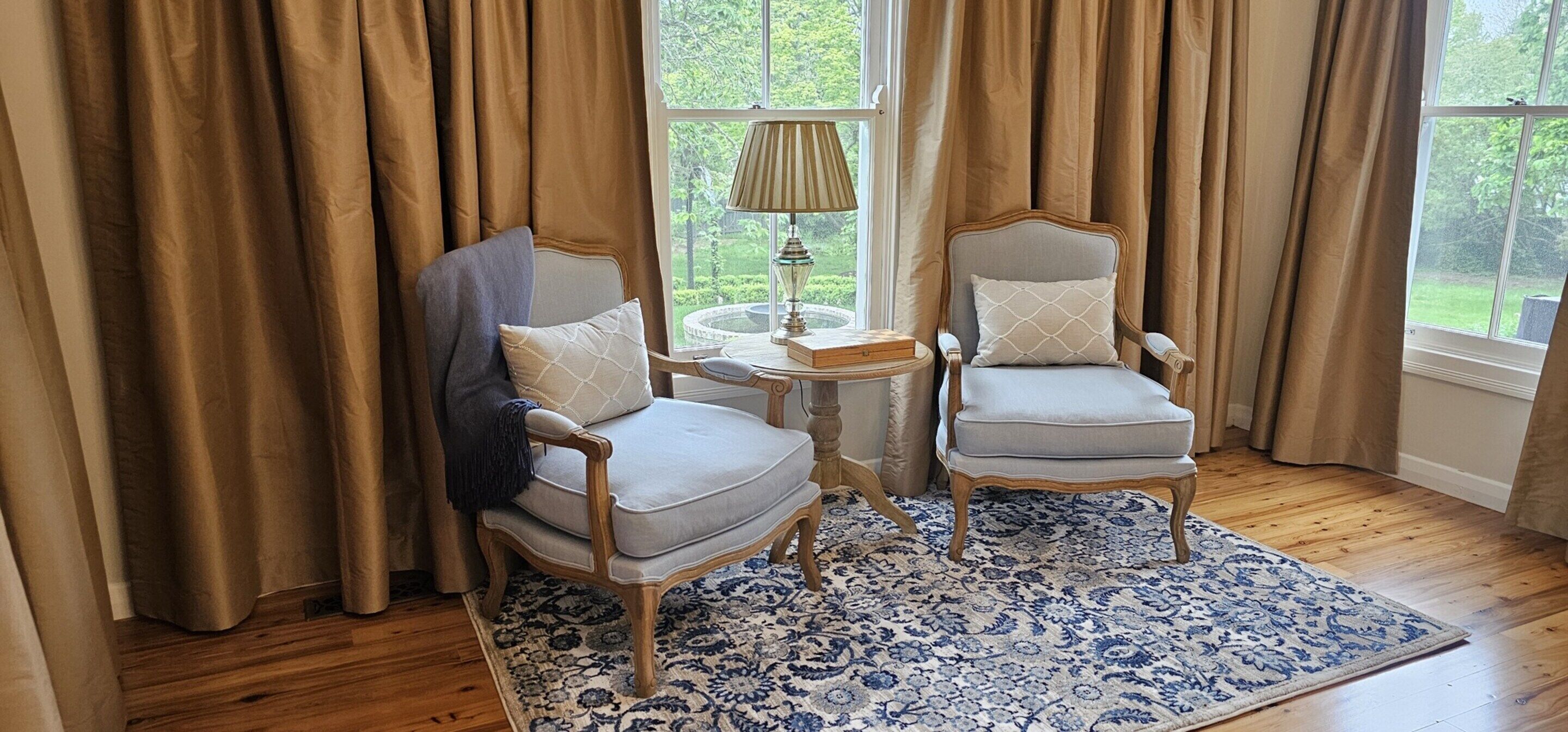 A room with two armchairs, a small round table, a lamp, and a view through a window with curtains.