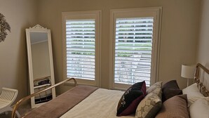 Un dormitorio con cama, una silla, un espejo y dos ventanas con persianas.