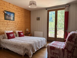 A bedroom with a wooden wall, a bed with a white and red bedspread, a chair, and a window with a view of greenery. - Cozy apartment with balcony - 5 guests (Villard-de-Lans)