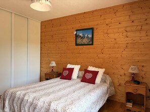 A bedroom with a wooden wall, a bed with a white textured blanket, two red pillows, a framed picture of mountains, and two bedside tables with lamps. - Cozy apartment with balcony - 5 guests (Villard-de-Lans)