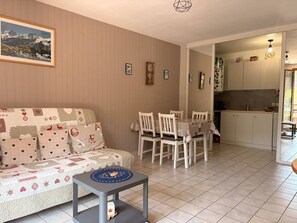 A living room with a sofa, a dining table with chairs, a small side table, and a kitchen area with cabinets and a sink. - Cozy apartment with balcony - 5 guests (Villard-de-Lans)