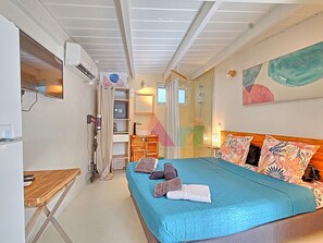 A bedroom with a bed, a wooden side table, a built-in wardrobe, a shower area, and a painting on the wall. - Ideal studio near beach, AC & parking (Saint-Martin)