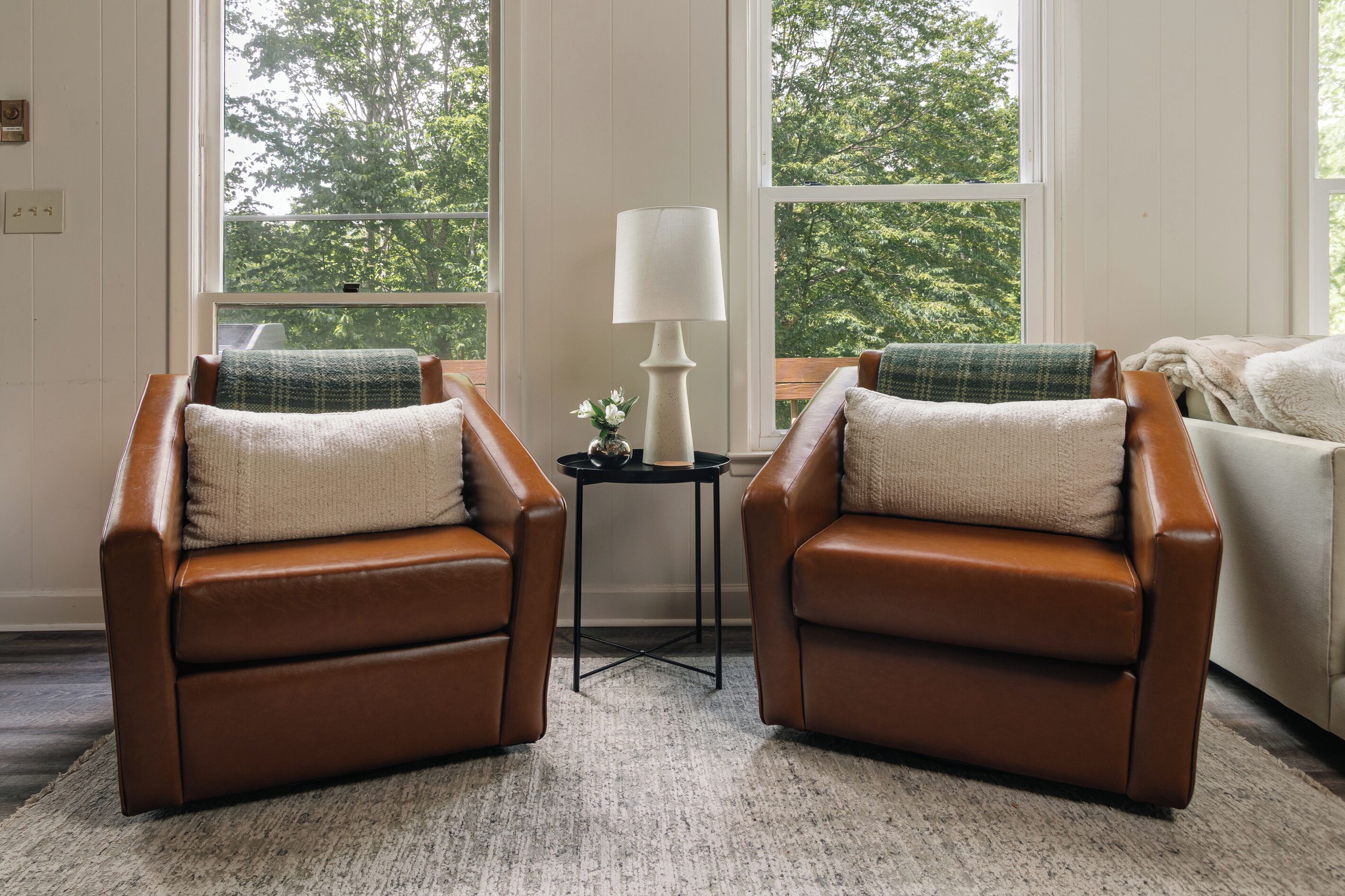 A room with two brown leather armchairs, a small table with a lamp, and a view of trees through large windows.