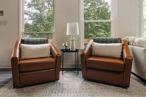 A room with two brown leather armchairs, a small table with a lamp, and a view of trees through large windows.