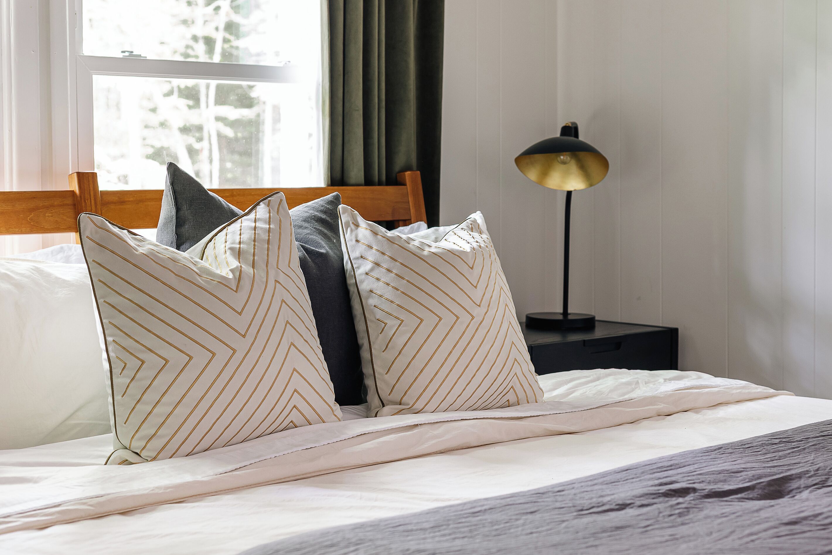 A neatly made bed with patterned pillows, a wooden headboard, a bedside lamp, and a window with curtains.