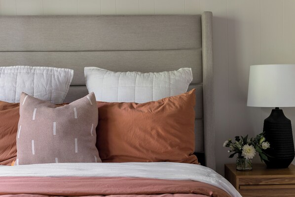 A neatly made bed with a grey headboard, multiple pillows in various colors and patterns, and a bedside table with a lamp and a vase of flowers.