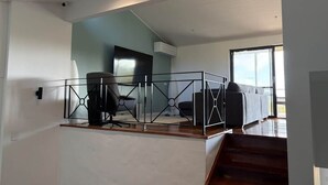 Een modern interieur met een trap naar een balkon, een zithoek met een bank en fauteuils, en een groot raam dat natuurlijk licht toelaat.