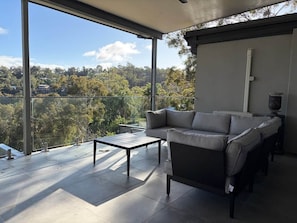 A modern balcony with a sofa and a table, offering a view of a green landscape. - Perth Hills Retreat | City Views | Luxury Beds (Gooseberry Hill)