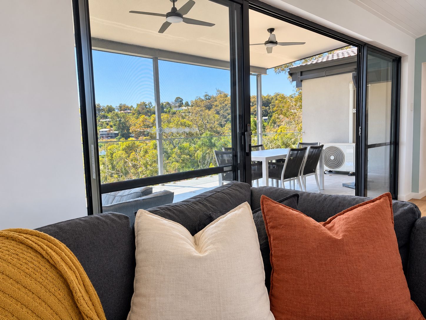 A modern living room with a sofa, a table, and chairs. There are large windows with a view of trees and a building.