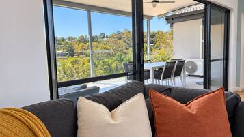A modern living room with a sofa, a table, and chairs. There are large windows with a view of trees and a building.