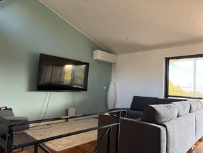 A modern living room with a flat-screen TV mounted on the wall, a grey sofa, and a large window providing a view.