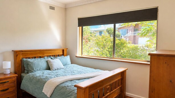 A bedroom with a wooden bed, a nightstand, a dresser, and a window with a view of greenery and buildings.